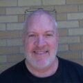 A smiling bald man with a beard, wearing a black t-shirt and glasses perched on his head, standing in front of a brick wall.