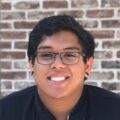 A smiling young man with glasses, wearing a black shirt, stands in front of a brick wall.