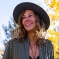 A smiling woman wearing a black hat and a green shirt stands outdoors with bright yellow autumn leaves in the background.