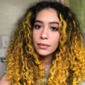 A close-up portrait of a young woman with vibrant, curly, yellow-dyed hair and a calm expression. She is looking directly at the camera, her face illuminated by soft light.