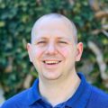 A smiling person wearing a blue shirt stands in front of a blurred leafy background. They are bald and looking directly at the camera.