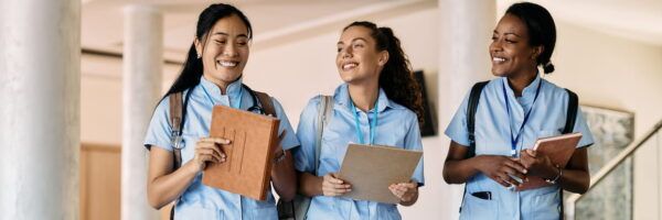 Three female nurses in scrubs, smiling and conversing while holding clipboards and notebooks in a hospital corridor, are majoring in Allied Health.