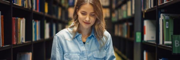 A college student in a denim shirt browses books in a library aisle, focusing intently on a selection. Shelves filled with colorful books line the background.