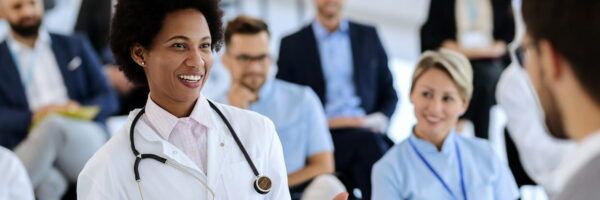 A diverse group of medical professionals in a seminar, including a black female nurse with a stethoscope around her neck, smiling and engaged in a discussion with colleagues about the nurse licensure compact.