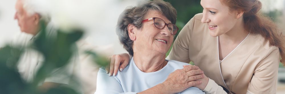 A young woman with red hair comforting an elderly woman wearing glasses, both smiling warmly in a softly blurred indoor setting.