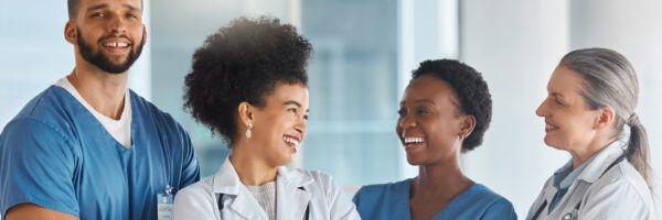 Four diverse healthcare professionals, including RNs and LVNs, smiling and interacting in a bright hospital corridor. Two are in scrubs, and two are in white coats.