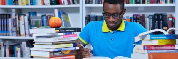 A focused young man wearing glasses and a blue polo shirt studies intensely among piles of books in a library, with a red apple beside him.