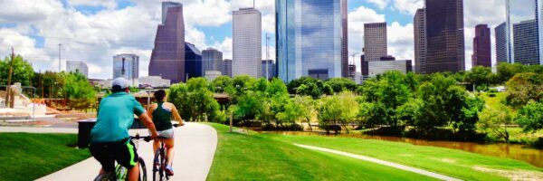 Two cyclists riding on a pathway beside a river, with a backdrop of a vibrant city skyline and clear blue sky. Lush greenery lines the riverbank under a sunny sky. Nearby, banners for