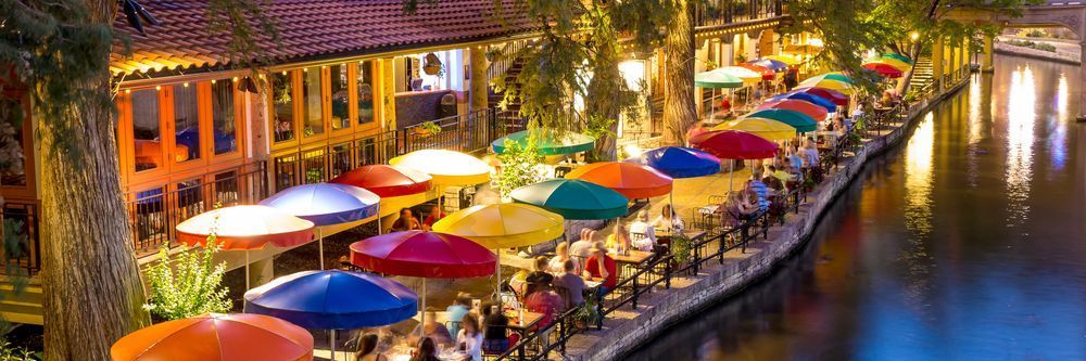 A bustling riverside dining scene at night, featuring colorful umbrellas and illuminated buildings reflecting on the water, with patrons enjoying meals.