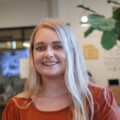 A woman with long blonde hair and a rust-colored top smiles at the camera. She is indoors, with blurred background elements including a large plant and wall decorations.