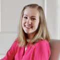 A person with medium-length blonde hair, wearing a bright pink blouse, sits on a light-colored chair and smiles warmly at the camera. The background is a neutral indoor setting, possibly a home or office.