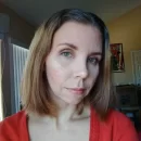 A woman with shoulder-length light brown hair and light skin is looking towards the camera. She is wearing a deep red V-neck shirt and is inside a room with soft natural light. There are framed pictures and some boxes in the background.