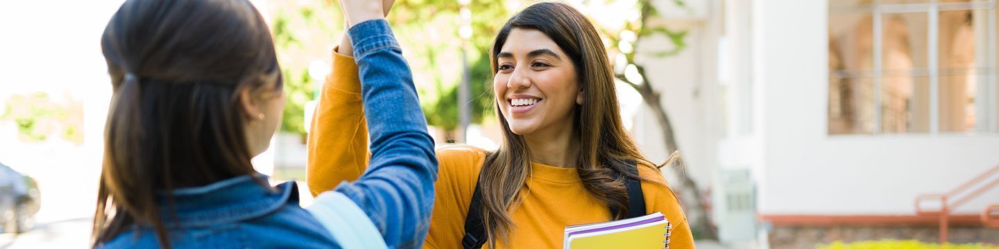 Two people greet each other enthusiastically outside on a sunny day. One, wearing a yellow sweater, holds notebooks and smiles while the other, in a blue jacket, raises a hand in greeting. As Latino nursing students, they share stories of their studies at one of the best online schools. Blurred trees and buildings are in the background.