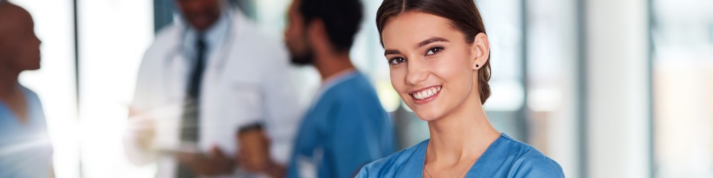 A smiling healthcare worker in blue scrubs, possibly a medical assistant, stands in the foreground while three students in medical attire converse in the blurred background. The scene is set in a bright, modern facility that inspires aspiring careers.