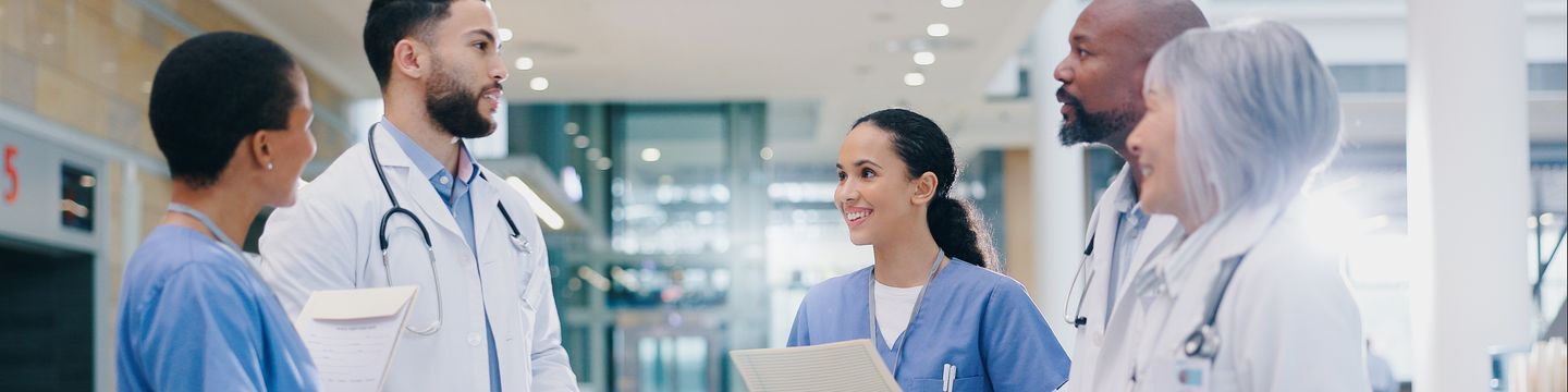 A diverse group of medical professionals, including a dedicated medical assistant, in scrubs and lab coats stand together, holding folders and engaging in a discussion about new opportunities in a bright hospital hallway.