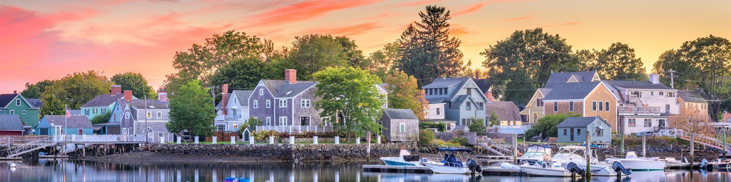 A picturesque waterfront village at sunset, featuring a row of quaint houses with various architectural styles. Boats are docked along the calm water, and trees add greenery to the serene scene. The sky is painted with soft pink and orange hues, reflecting tranquility perfect for unwinding from coding programs.