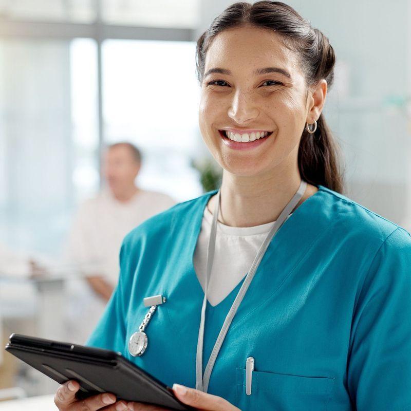 A group of four healthcare professionals in blue scrubs engaging in a discussion while looking at a digital tablet, sitting together in a room with brick walls.