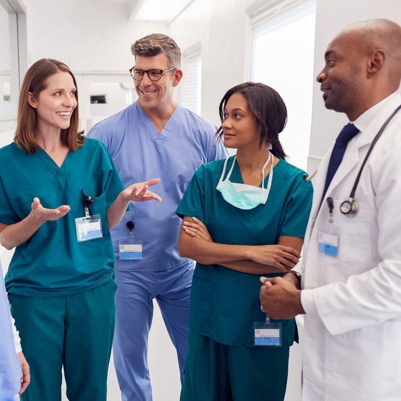 A diverse group of bilingual medical professionals in scrubs, including men and women, applauding during a meeting in a bright clinical setting.