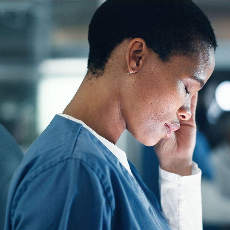 A female nurse in a white lab coat looks stressed or fatigued, sitting at her desk with her hands on her forehead, a laptop open in front of her.