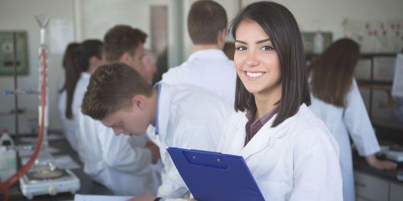 A focused male scientist in a blue lab coat examines samples through a microscope in a high-tech lab, with a female colleague working in the background.