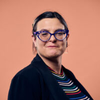 Against a peach background, a person with short dark hair smiles slightly, wearing blue cat-eye glasses and dangling triangular earrings. Their ensemble features a striped shirt and a black blazer, adding a touch of sophistication to their chic look.